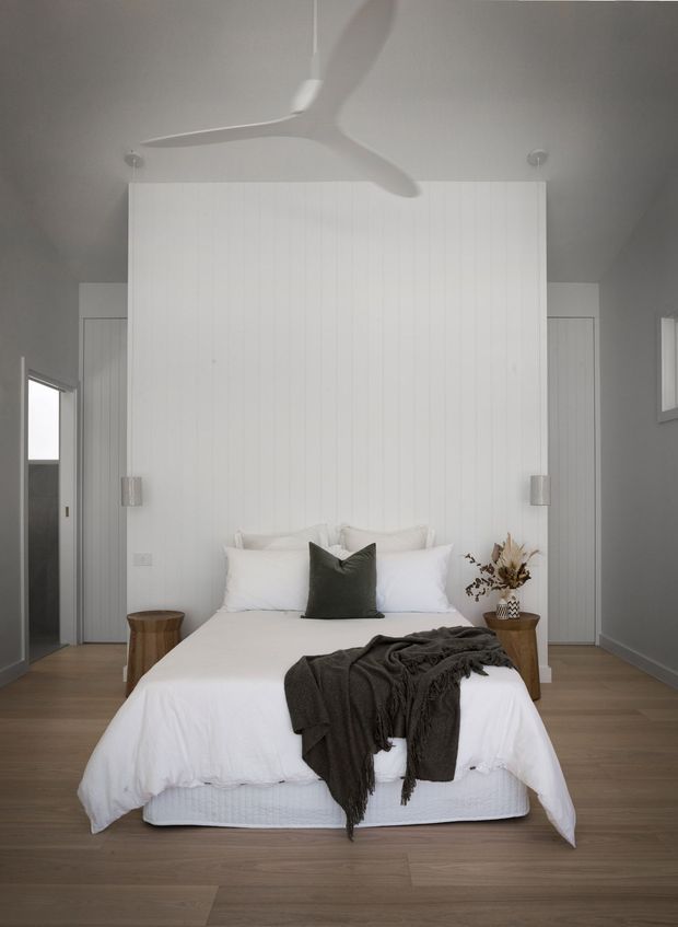 A serene, minimalist bedroom with a centered upholstered bed dressed in white bedding, a dark charcoal throw and cushion, and small timber bedside tables. The room features a full-height white vertical-panel feature wall behind the bed, concealed or flush wardrobe doors, pale grey walls, warm timber flooring, soft wall-mounted lights, and a sculptural white ceiling fan.
