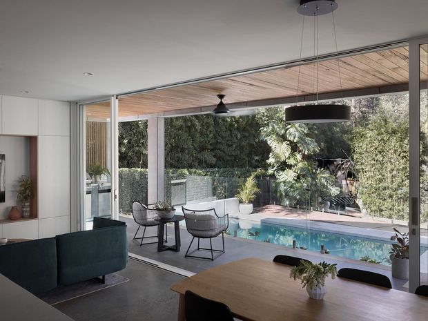 A bright, contemporary open-plan living and dining area opening directly onto a covered outdoor terrace and swimming pool. Floor-to-ceiling sliding glass doors create a strong indoor–outdoor connection. The interior features a dark teal sofa, timber dining table, white built-in cabinetry, polished concrete flooring, and a timber-lined ceiling extending across the terrace. Dense garden screening surrounds the pool outside.