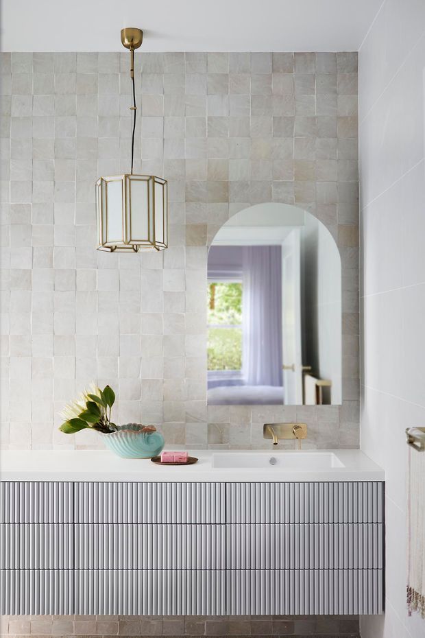 A refined bathroom vanity with a long ribbed grey built-in cabinet, white integrated basin, pale textured square wall tiles, and brushed brass tapware. A geometric brass pendant hangs above the vanity, while an arched mirror reflects a softly coloured adjacent room. The palette is light and understated with subtle decorative styling.