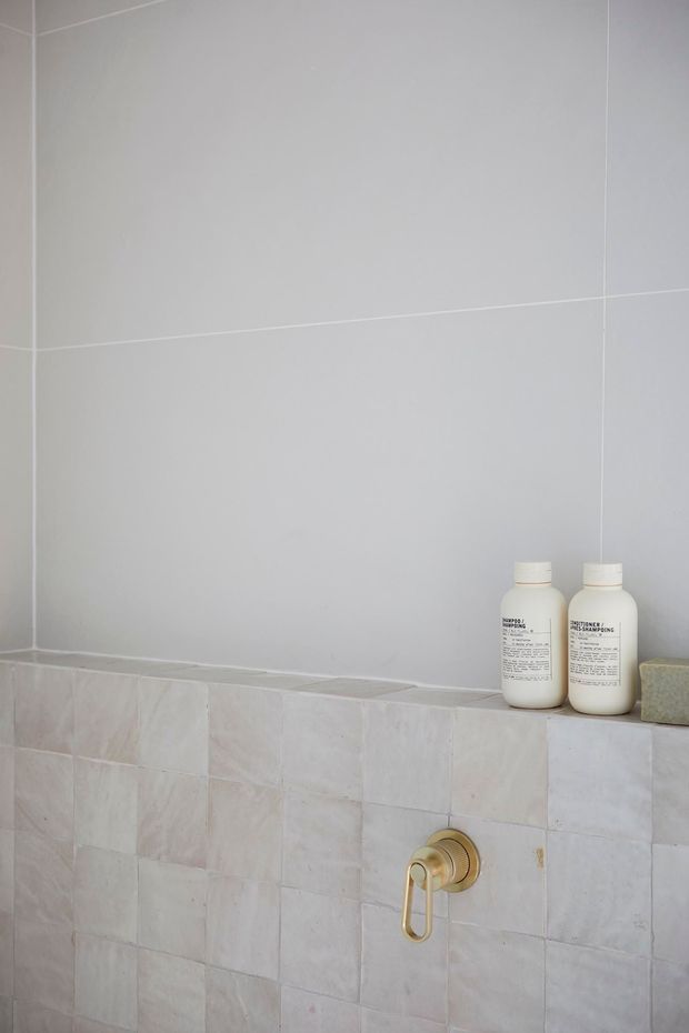 Close-up of a minimalist bathroom wall featuring large light-grey tiles above smaller pale stone-look tiles, with a recessed ledge holding two white bottles and a gold-toned wall-mounted tap control. The scene is clean and softly lit, with a neutral palette and subtle material contrast.