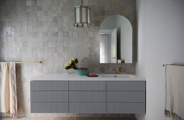 Refined contemporary bathroom featuring a wall-mounted, vertically fluted grey vanity with an integrated white basin, a large arched mirror, pale textured stone-look wall tiles, and brass tapware and towel rails. Soft textiles, a sculptural pendant light, and a small plant add warmth to the otherwise calm neutral scheme.