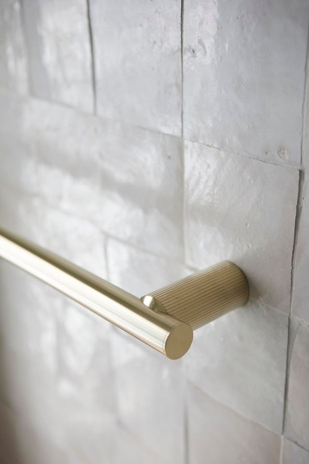 Close-up detail of a brushed brass or gold-toned cylindrical bathroom towel rail mounted on a textured white rectangular tile wall. The ribbed cylindrical wall bracket and softly reflective metal finish create a refined, minimal fixture detail.