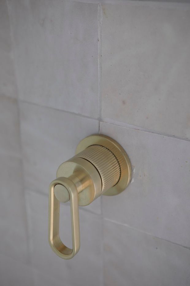 Close-up of a brushed brass/gold-toned wall-mounted shower control with a ribbed cylindrical body and elongated lever, set into large pale stone or ceramic wall tiles with visible grout joints.