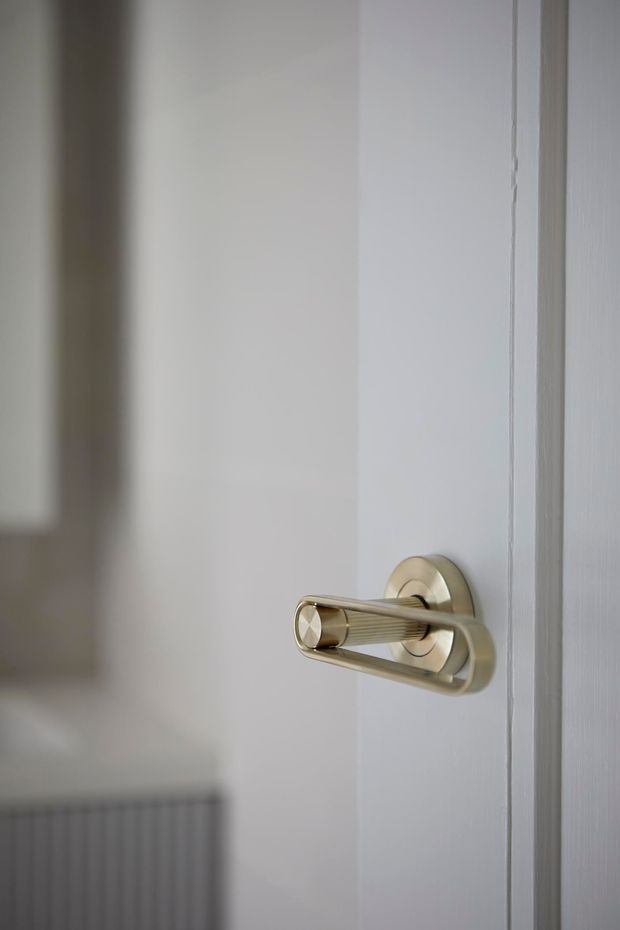Close-up interior photograph of a white painted panel door with a distinctive brushed brass or gold-toned lever handle and circular backplate. A softly blurred white radiator and neutral wall are visible in the background.