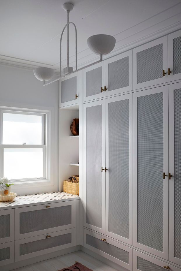 A bright, compact built-in storage room with floor-to-ceiling pale cabinetry, woven or mesh inset door panels, brass pulls, and matching drawer units beneath a window. A patterned countertop runs below the window, with open shelving holding baskets and decorative items. The room has light painted walls, a white ceiling, pale flooring, and a sculptural linear pendant light.
