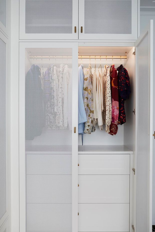 Close-up view of a built-in white wardrobe with mesh-front cabinet doors, brass-toned handles and hanging rail, displaying clothing above integrated drawers. The cabinetry is brightly lit and neatly fitted into the wall, with a refined, traditional-inspired appearance.