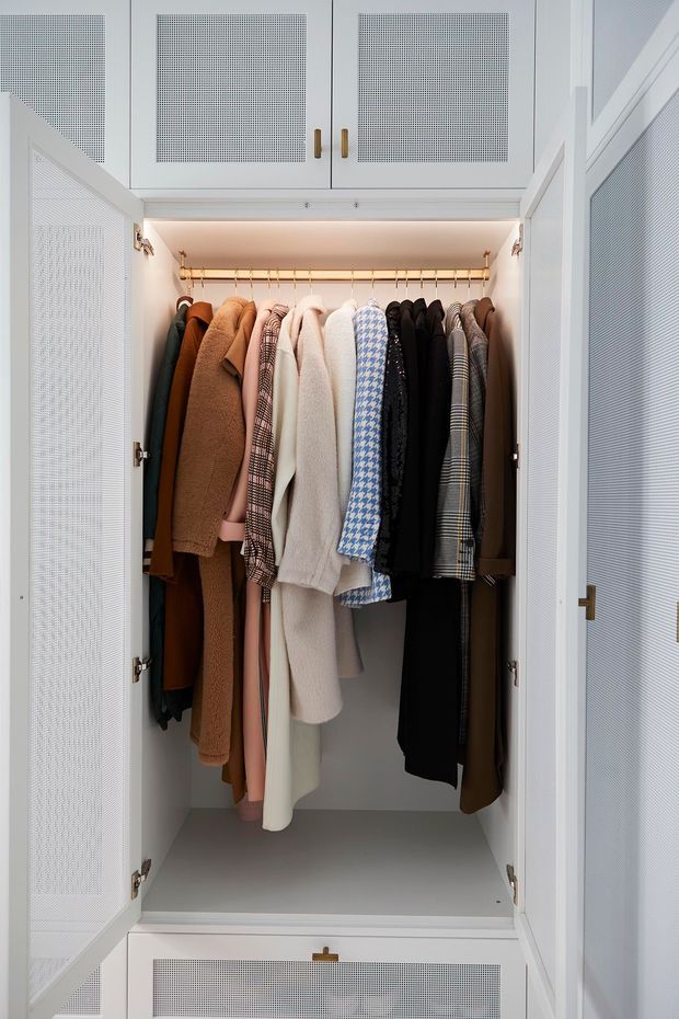 A close-up view of a built-in wardrobe cupboard with open white perforated-panel doors, a brass hanging rail, integrated warm lighting, and neatly arranged coats and garments. Matching overhead and lower cabinet doors frame the hanging compartment.