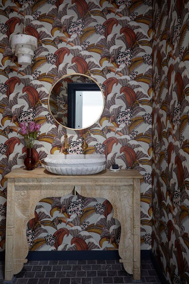 A richly patterned powder room with dramatic botanical wallpaper covering the walls, a round brass-framed mirror, sculptural white vessel basin, carved pale stone console, dark tiled floor, and a decorative pendant light. The space has an ornate, theatrical character with warm metallic and deep jewel-toned accents.