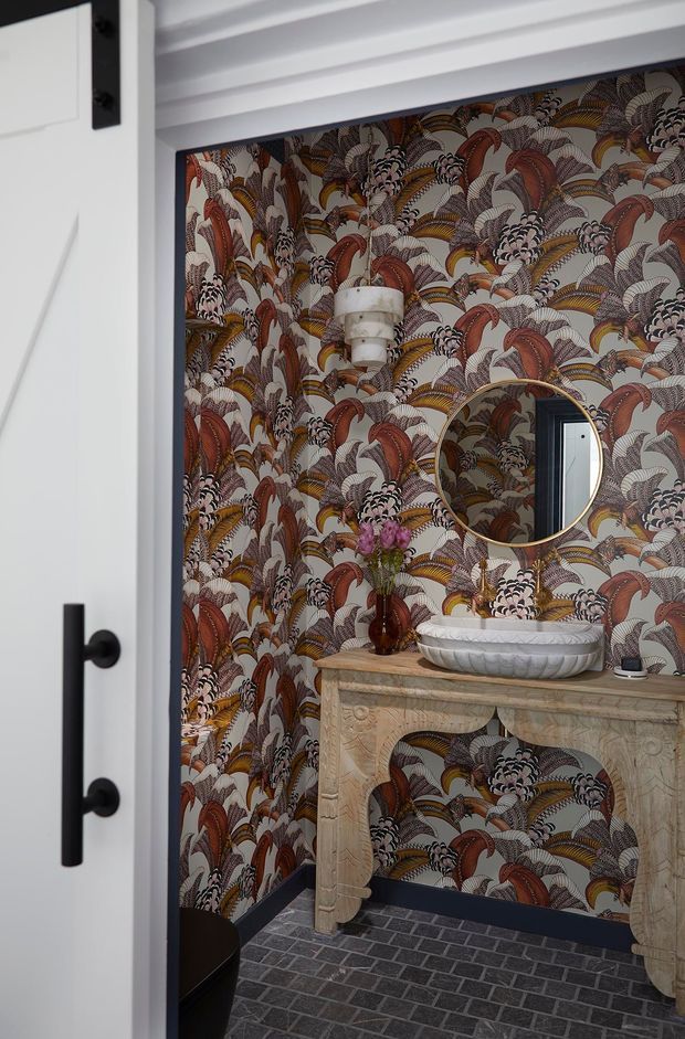 A compact, highly decorative powder room viewed through a partially open white door. The walls are covered in bold patterned botanical wallpaper in rust, cream, charcoal and muted gold tones. A carved light-stone console supports a sculptural white vessel basin, with a round brass-framed mirror and wall sconce above. The floor is finished in dark grey brick-pattern tiles, with black trim and hardware adding contrast.