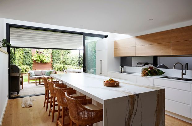 Bright contemporary kitchen with a large white stone island and integrated waterfall end, seating for several people on woven timber stools, white cabinetry, and extensive warm timber overhead cabinetry. Full-height black-framed glazing opens toward a leafy outdoor courtyard or patio, while a small adjoining outdoor seating area is visible beyond.
