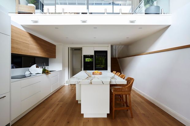 Bright contemporary kitchen and dining area with white handleless cabinetry, a warm timber overhead cabinet, marble-look waterfall island, integrated appliances, timber flooring, and woven timber bar stools. A loft-like mezzanine or upper-level opening with glass balustrading is visible above the kitchen, adding height and natural light.