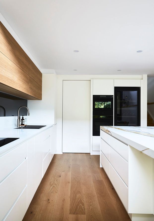 Bright contemporary galley kitchen with white flat-panel cabinetry, integrated black appliances, a white stone island benchtop, timber wall cabinetry, and warm timber flooring. Recessed ceiling downlights and minimalist detailing create a clean, streamlined interior.