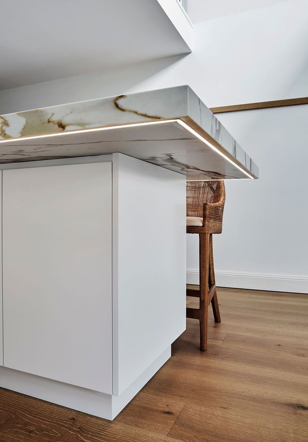 Close-up interior view of a contemporary kitchen island with seamless white cabinetry and a dramatic white veined stone benchtop featuring integrated linear underlighting. Warm timber flooring and a woven timber bar stool add contrast, with a bright minimalist interior and partial ceiling detail visible.