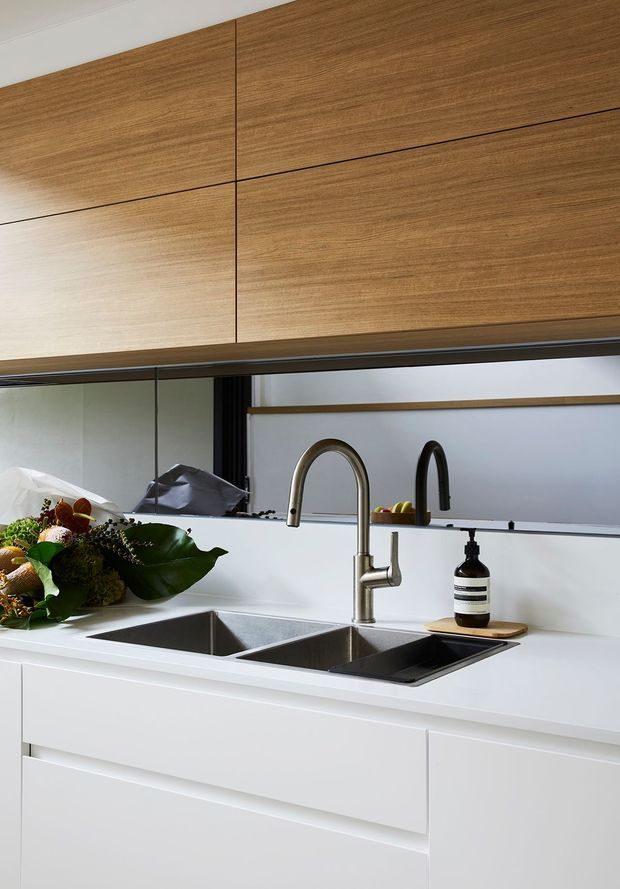 Close-up view of a contemporary kitchen sink area featuring white cabinetry and benchtops, a large stainless-steel double-bowl undermount sink, dark brushed-metal gooseneck and secondary tapware, and warm timber-grain overhead cabinetry. A mirrored splashback reflects the opposite side of the kitchen, while a narrow horizontal window or glazed opening provides additional light.