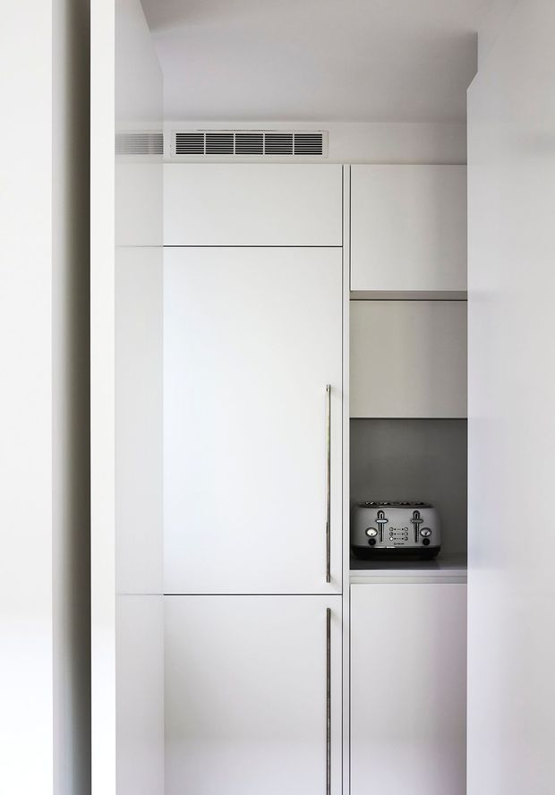 A compact, minimalist built-in kitchen or kitchenette concealed within a tall white cabinetry wall. The cabinetry includes a large integrated appliance or pantry door, overhead storage, open shelving with a small countertop appliance, and a dark grey backsplash or rear panel. The space is bright, monochromatic, and highly streamlined, with slim vertical metal handles and a linear ventilation grille above.