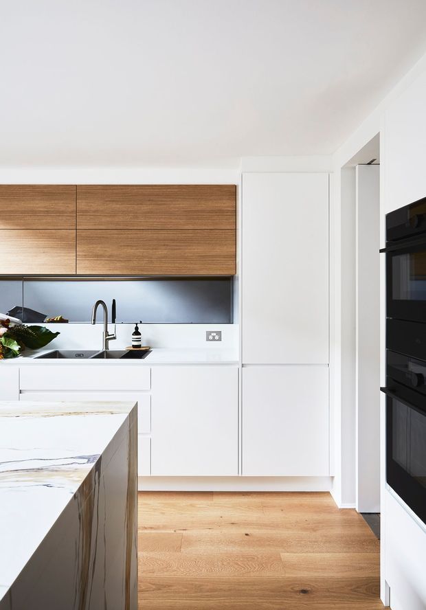 A bright, contemporary kitchen with white flat-panel cabinetry, warm horizontal timber overhead cupboards, a pale veined stone island benchtop, stainless-steel sink and tap, dark reflective splashback, integrated tall cabinetry, and stacked black appliances. Light timber flooring and a predominantly white palette create a clean, refined look.