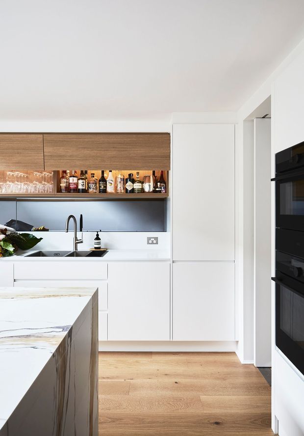 Bright contemporary kitchen with white flat-panel cabinetry, a timber-toned overhead cabinet and open drinks shelf, stainless steel sink and tap, dark reflective splashback, integrated black ovens, pale timber flooring, and a large white marble-look island with prominent veining.