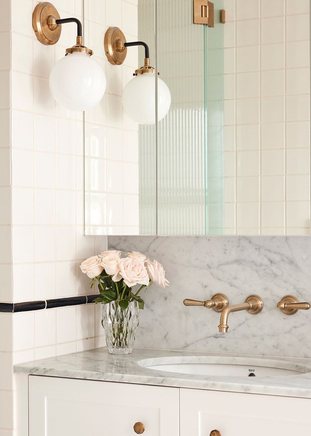 Close-up of a refined bathroom vanity featuring white square wall tiles with a black horizontal trim, a marble backsplash and countertop, white cabinetry, brass wall-mounted tapware, and matching brass globe wall sconces. A large mirror with a fluted glass panel detail is above the basin, with a vase of pale roses on the vanity.