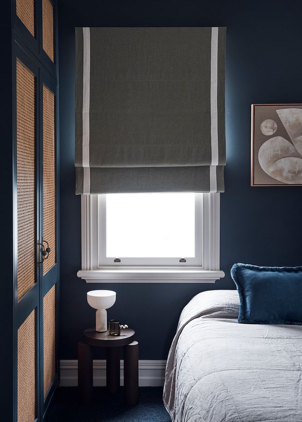 A moody, refined bedroom corner with deep navy walls, white traditional window trim, and a grey Roman shade over the window. The room includes a partially visible bed dressed in white textured bedding with a navy velvet cushion, a dark round bedside table with a small lamp, woven-panel navy cabinetry or wardrobe doors, and abstract wall art.