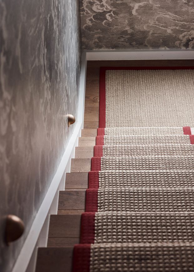 A close-up view looking down a narrow stairway with timber stair treads, a textured patterned wall, and a woven stair runner bordered in deep red. The runner continues onto the upper landing, creating a warm, richly layered and tactile interior. Small rounded timber handrail supports are visible along the wall.