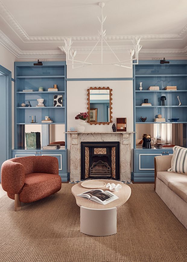 A refined living room centered on a decorative fireplace with a marble surround and ornate dark firebox. Symmetrical blue built-in shelving and cabinetry flank the fireplace, displaying books and objects. The room features elaborate white ceiling cornices, a sculptural white pendant light, a scalloped wood-framed mirror, a rust-red upholstered armchair, a neutral sofa, and a rounded coffee table over a textured brown carpet.