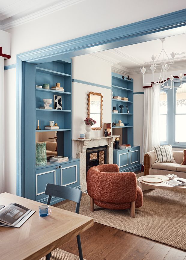 A refined living room with blue built-in shelving and cabinetry framing a decorative marble fireplace. The room features white walls with blue trim, ornate ceiling detailing, a sculptural chandelier, warm timber flooring, a neutral rug, a rust-coloured lounge chair, and a beige sofa. A timber table and blue dining-style chair occupy the foreground, while tall windows provide natural light.