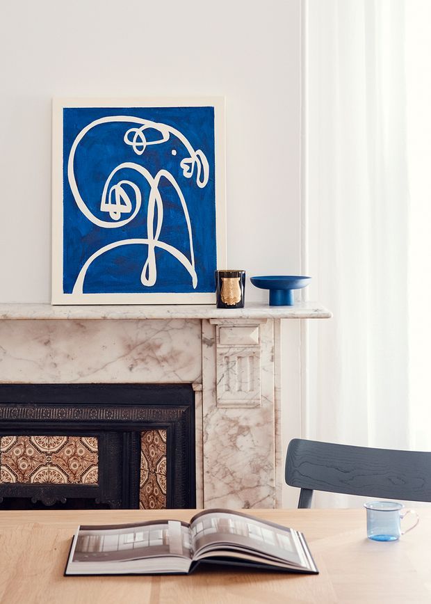 A refined interior vignette centred on an ornate marble fireplace with a dark decorative surround and patterned tiled insert. A blue abstract artwork, blue ceramic bowl, and small decorative vessel sit on the mantel, while a timber dining table with an open book and blue glass occupies the foreground. Soft daylight filters through sheer curtains beside a dark timber chair.