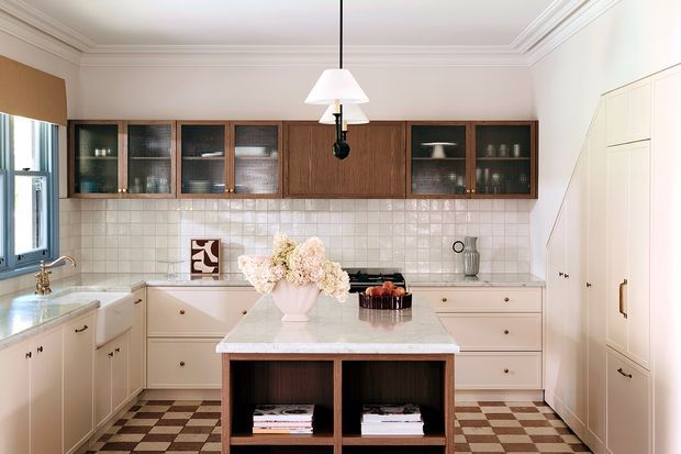 A refined traditional kitchen with cream-painted cabinetry, dark timber-framed upper display cabinets, a pale stone island, and a checkerboard tile floor. Decorative cornices, brass-toned hardware, a tiled splashback, and a pendant light contribute to the classic, warm character.