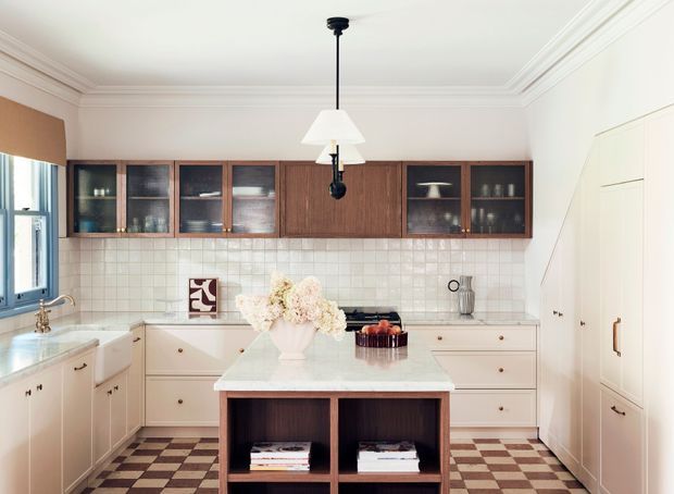 Bright traditional kitchen with cream cabinetry, a central island, dark timber overhead display cabinets, white tiled splashback, checkerboard floor tiles, and a hanging pendant light. The room has high decorative ceiling cornices, integrated wall storage, and a softly refined, classic interior.