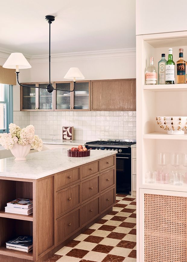 A warmly styled kitchen with a large central island featuring timber cabinetry and a pale stone benchtop. The room includes a checkered brown-and-cream floor, tiled splashback, vintage-style range cooker, glazed upper cabinets, open shelving with bottles and glassware, and a black linear pendant light. Soft pink and cream tones, timber joinery, and floral arrangements create a refined, inviting atmosphere.