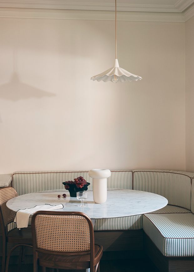 A refined dining nook with a curved built-in banquette upholstered in narrow green-and-cream stripes, surrounding a pale oval marble table. A cane-backed dark timber dining chair sits in the foreground, with a simple white pendant light above and a small floral arrangement on the table. The room features warm cream walls and ornate ceiling cornices, creating a soft, elegant atmosphere.