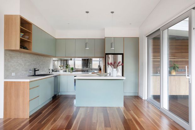 Bright contemporary kitchen with pale blue-green flat-panel cabinetry, warm timber storage accents, a central island, light stone-look splashback, dark timber flooring, and large sliding glass doors opening to a timber deck. The space has clean lines, integrated appliances, and abundant natural light.