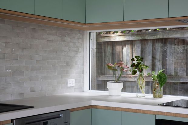 Compact contemporary kitchen corner with muted sage-green flat-panel cabinetry, warm timber trim, a pale engineered-stone benchtop, and a light grey textured masonry-tile splashback. A wide horizontal window brings in natural light and overlooks a timber fence, with potted plants arranged on the sill. Integrated appliances and a cooktop are visible below the benchtop.