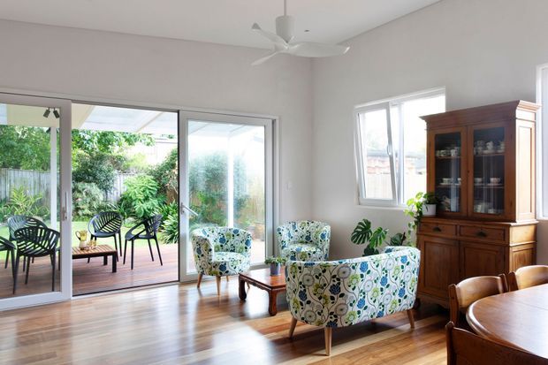 Bright living room with polished timber flooring, pale walls, a white ceiling fan, large sliding glass doors opening to a leafy outdoor area, patterned upholstered armchairs and sofa, a wooden coffee table, and a traditional timber display cabinet. A dining table and chairs are partially visible in the foreground.