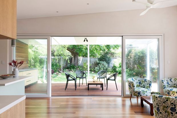 Bright contemporary open-plan living area with warm timber flooring, pale walls, a partial kitchen at left, and wide sliding glass doors opening to a timber deck and landscaped garden. Patterned upholstered armchairs frame the foreground, while outdoor seating is visible beyond the glazing. A ceiling fan and recessed lighting complete the airy interior.