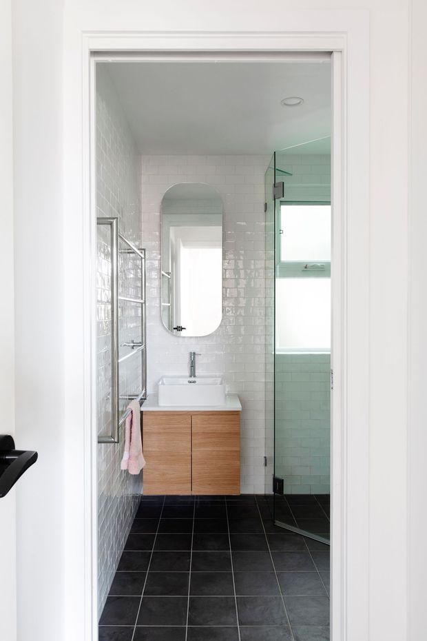 Bright, narrow contemporary bathroom viewed through a white-trimmed doorway. The room features glossy white rectangular wall tiles, dark charcoal floor tiles, a floating timber vanity with a white basin, rounded-rectangle mirror, wall-mounted fixtures, and a frameless glass shower enclosure with a tiled shower area and window.