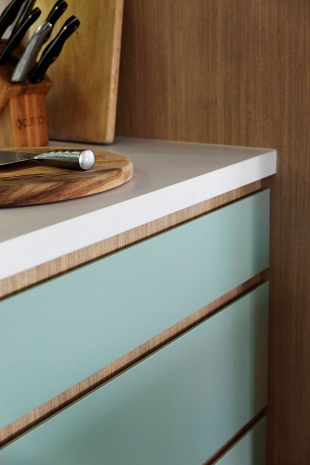 Close-up detail of a contemporary kitchen cabinet or built-in storage unit. The cabinetry has muted sage-green drawer fronts framed by warm wood-grain edging, a crisp white benchtop, and a darker timber wall or panel behind it. A wooden cutting board, knife block, and kitchen knife sit on the benchtop, emphasizing the kitchen setting.