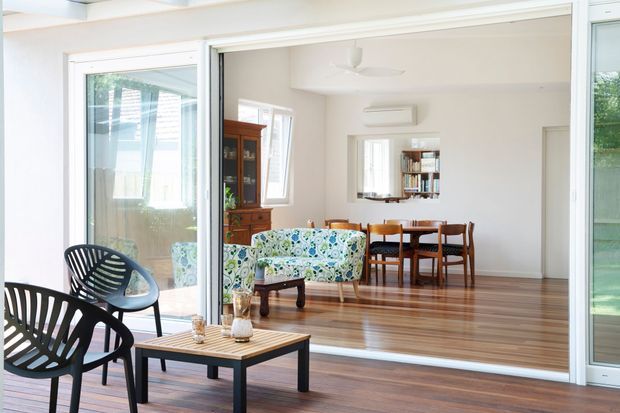 Bright open-plan living and dining area viewed from a glazed outdoor-facing space. The room features polished timber flooring, white walls and ceiling, broad sliding glass doors, a patterned floral sofa, dark modern lounge chairs, a low coffee table, timber dining furniture, and a traditional wooden display cabinet. The interior is airy and connected to the outdoors.