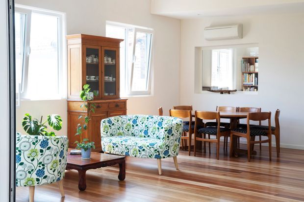 Bright open-plan living and dining room with polished timber flooring, white walls, large windows, patterned upholstered armchairs and sofa, a wooden display cabinet, a coffee table, and a traditional timber dining set. A wall-mounted air conditioner, indoor plants, and a bookshelf are also visible.