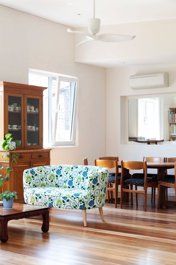 Bright open-plan living and dining area with polished timber flooring, pale walls, a white ceiling fan and wall-mounted air conditioner. A patterned floral loveseat and dark timber coffee table sit in the foreground, while a traditional timber china cabinet, dining table and wooden chairs occupy the background. A large interior opening provides a view into an adjacent room with shelving.