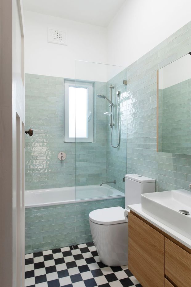 Bright compact bathroom with pale mint-green glazed subway wall tiles, a black-and-white checkerboard floor, white bathtub with clear glass shower screen, wall-mounted shower fixtures, white toilet, and a white rectangular basin on warm timber vanity cabinetry. A large mirror and narrow frosted window add light to the space.