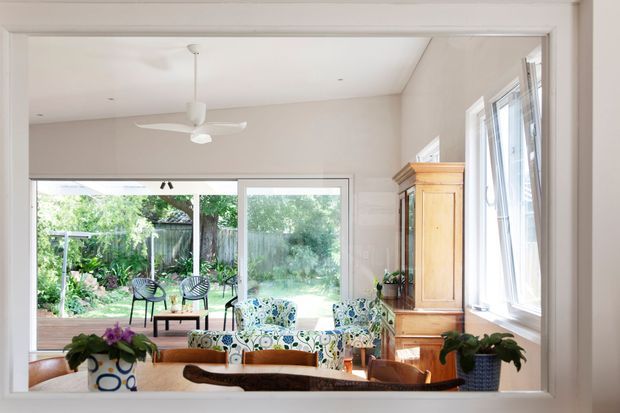 Bright, airy sunroom or living area viewed through a wide interior opening, with white walls and ceiling, large windows and sliding glass doors opening to a leafy garden, patterned upholstered armchairs, timber furniture, potted plants, and a ceiling fan. The room has a relaxed traditional-contemporary character and abundant natural light.