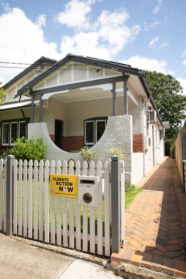 Bright exterior view of a single-storey heritage-style brick-and-render house viewed from the street. The facade features a covered front porch with timber posts, decorative curved rendered privacy walls, white-framed windows, a dark pitched gabled roof, and a white picket fence with a pedestrian gate. A narrow brick-paved side path runs alongside the house.