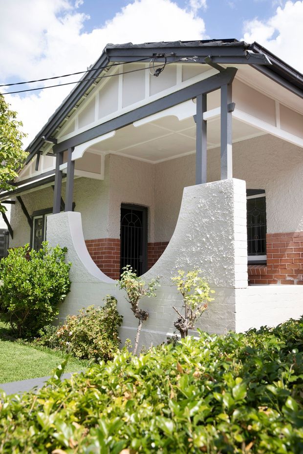 Exterior view of a single-storey period bungalow with a covered front verandah, white textured rendered walls, dark grey timber framing and gable detailing, red brick dado, and decorative curved verandah privacy walls. Mature hedges and garden planting frame the foreground.