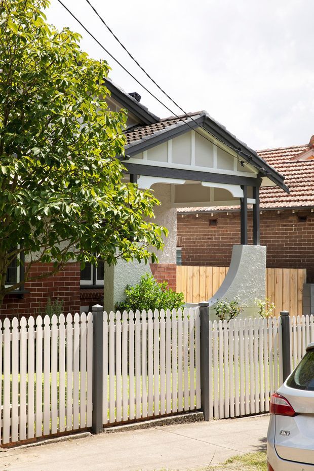 Exterior view of a traditional single-storey suburban house with a front porch, gabled roof, exposed decorative gable framing, red brick walls, rendered porch columns, and a white picket fence. Mature greenery partially screens the facade, with a driveway and neighbouring roof visible.