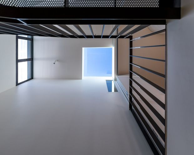 Upward-looking view of a minimalist contemporary interior void or stairwell, with white walls, tall glazing, a bright overhead opening, and dark horizontal metal balustrades and ceiling beams creating a strong geometric composition.