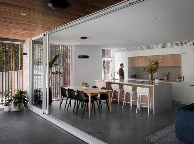 Open-plan contemporary kitchen, dining and living area viewed through wide glazed sliding doors. The space features a large grey island with white bar stools, timber-toned overhead cabinetry, a long timber dining table with black chairs, dark grey flooring, white walls, and a warm timber-lined ceiling extending toward the covered outdoor area. Vertical screened glazing and indoor plants add texture and connection to the exterior.