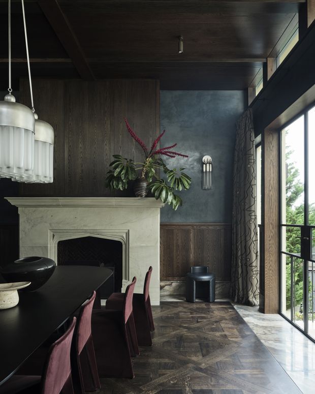 A moody, refined dining room with a dark timber ceiling and wall panelling, a pale stone fireplace, patterned dark timber flooring, and tall black-framed windows opening to greenery. A long black dining table with sculptural pendant lights and muted burgundy upholstered chairs occupies the foreground, while a small black stool, decorative wall light, and floral arrangement complete the richly layered interior.