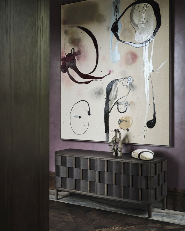 A moody interior vignette featuring a large abstract artwork in a dark frame above a sculptural dark timber console. The console has a textured, woven-block style facade and slim metal legs, with small sculptural objects and a rounded decorative bowl on top. Deep mauve walls, dark timber flooring in a herringbone pattern, dark vertical timber wall panelling, and subdued lighting create a refined, dramatic atmosphere.