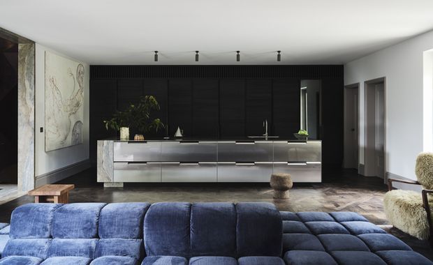 Contemporary open-plan living space with a dramatic black kitchen wall and long central island, viewed from behind a plush deep-blue sectional sofa. The kitchen features dark vertical ribbed cabinetry, a pale stone waterfall island, integrated appliances, and minimal black track lighting. Dark herringbone timber flooring, large artwork, indoor plants, and sculptural furniture create a refined, moody interior.